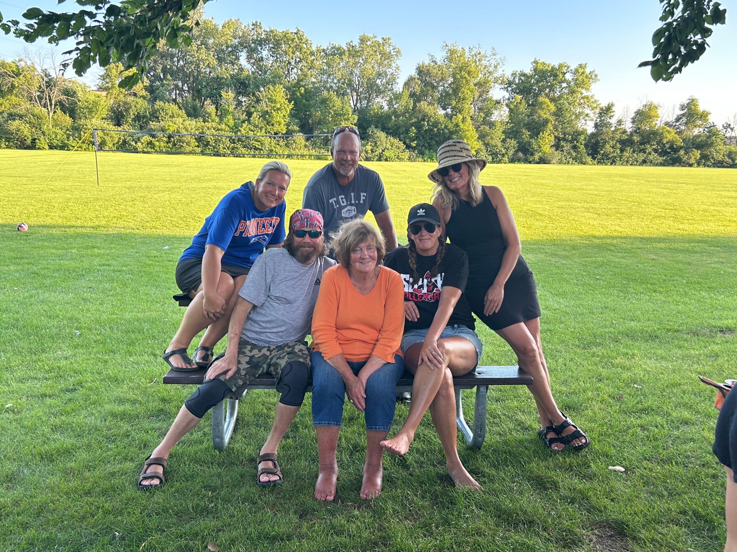 Group of six adults posing on a park bench on a sunny day, with a volleyball net and green field in the background.