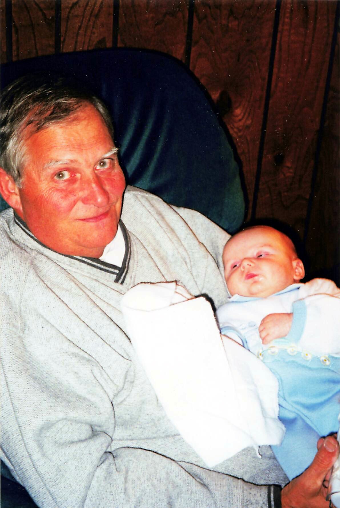 Grandfather in a light gray sweater gently holding a newborn baby wrapped in a white blanket with a blue outfit, against a wooden background.