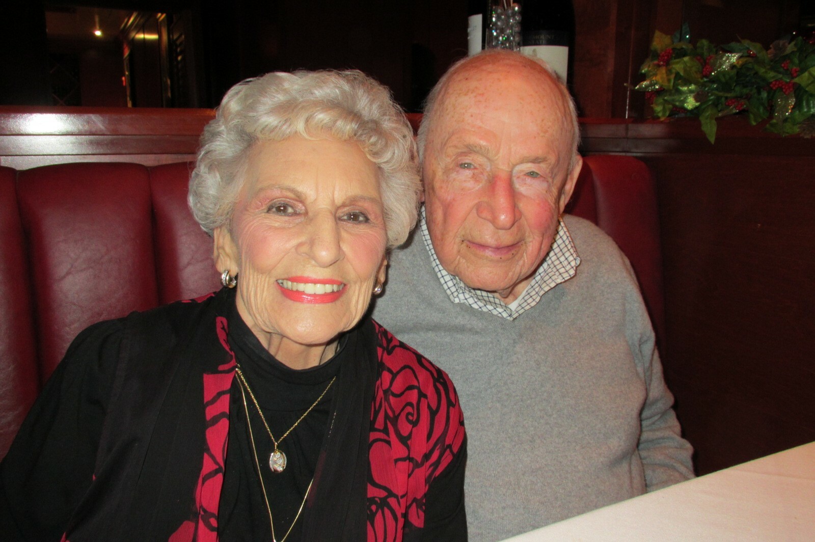 Smiling elderly couple seated in a red vinyl restaurant booth, the woman wearing a red and black patterned jacket.