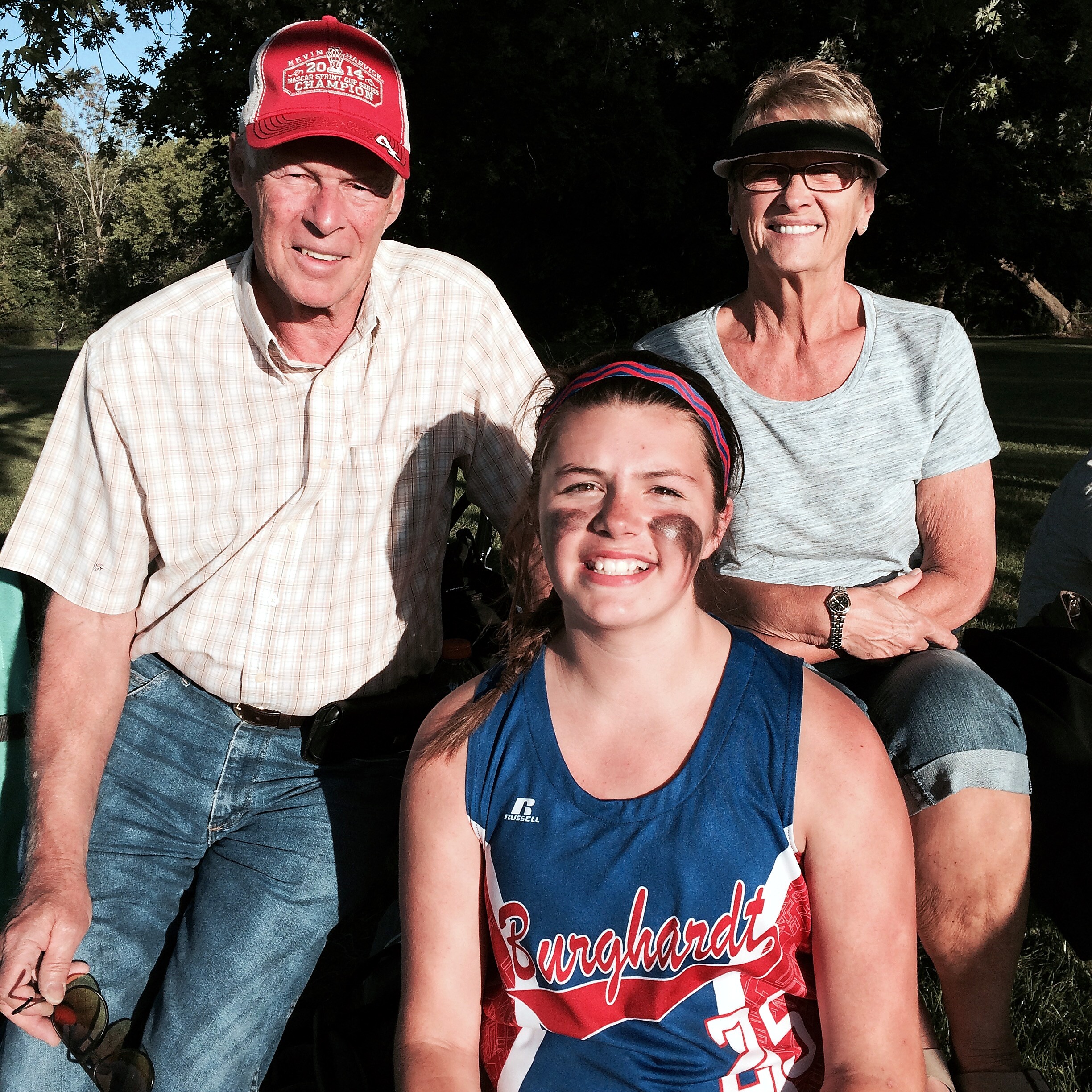 Three people posing outdoors on grass and trees, smiling at the camera (adult man with red cap, woman with visor, child in front wearing a blue sports jersey).