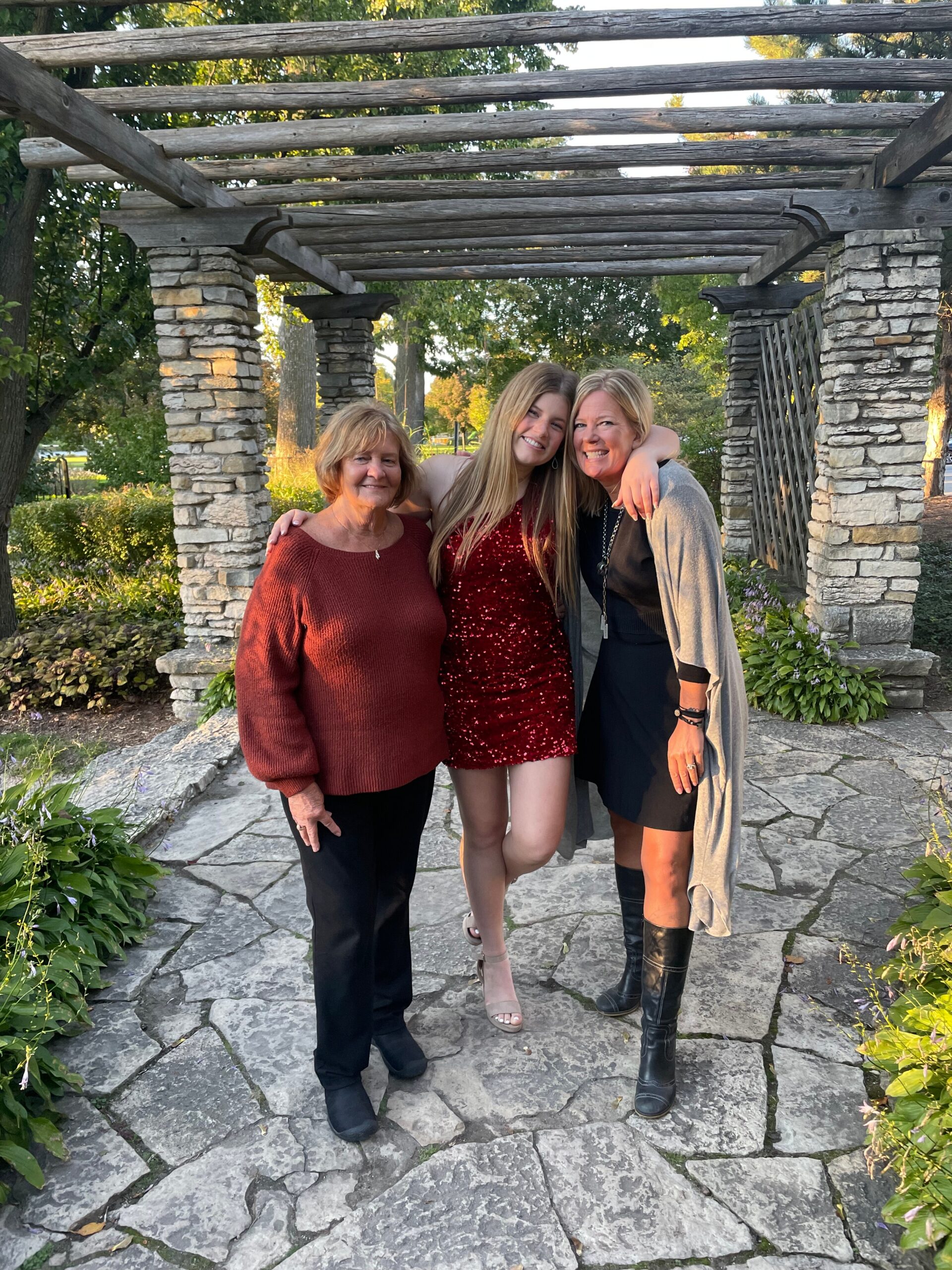 Three women posing and smiling under a rustic wooden pergola in a garden setting.