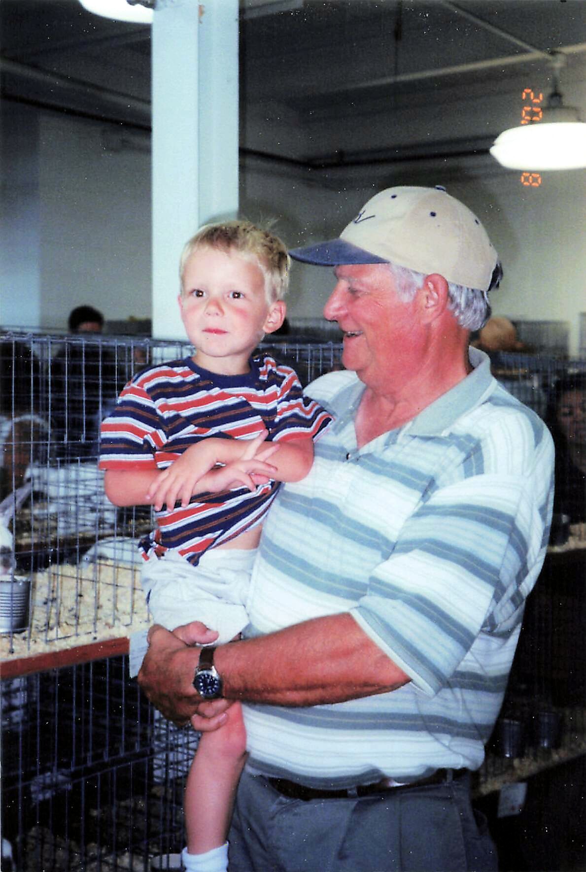 Grandfather in a cap holding a young boy in a striped shirt at an indoor pet show, cages in the background.