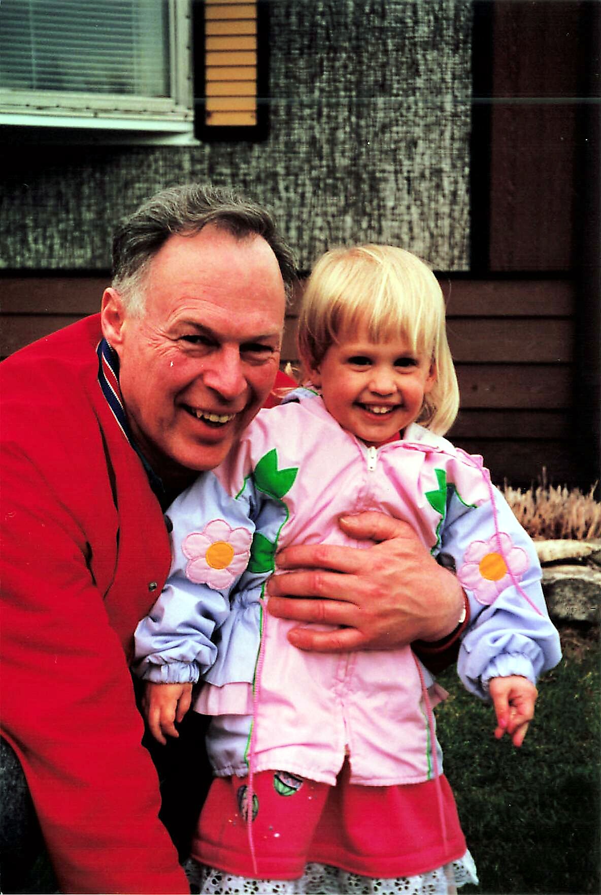 Older man in a red jacket hugs a smiling young girl in a pink floral jacket outdoors in front of a house.
