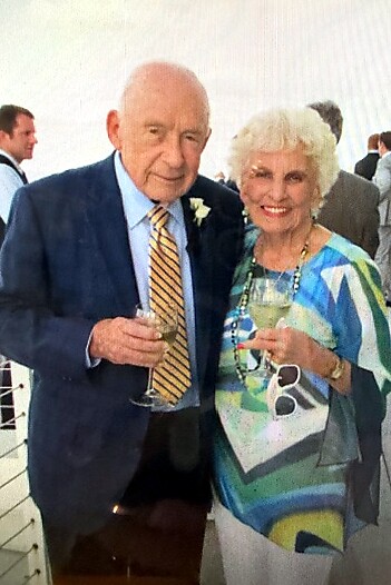 Smiling elderly couple with champagne glasses at a social event: man in a blue blazer and striped tie, woman in a colorful dress with sunglasses hanging, boutonniere on man.