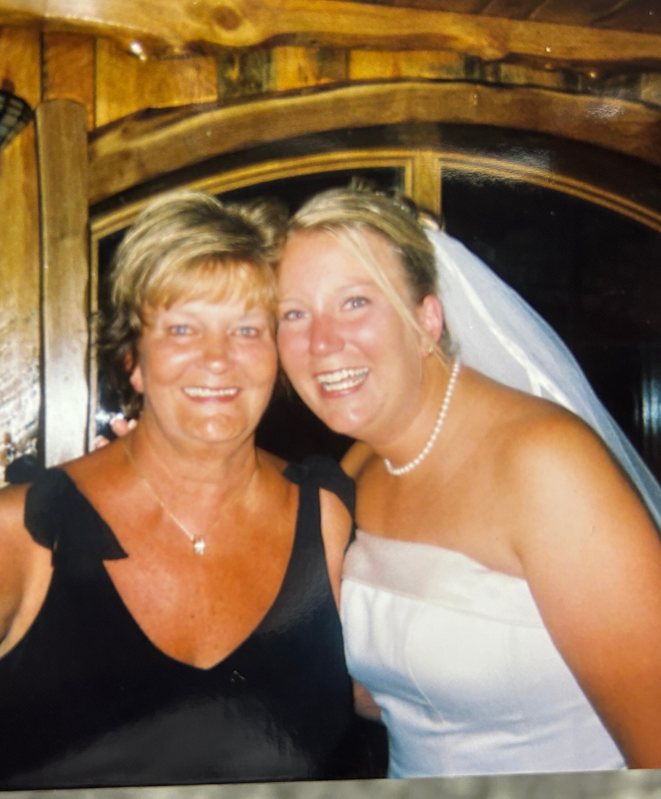 Two women posing and smiling for a photo, one in a black dress and the other in a white wedding gown with a veil, indoors against wooden decor.