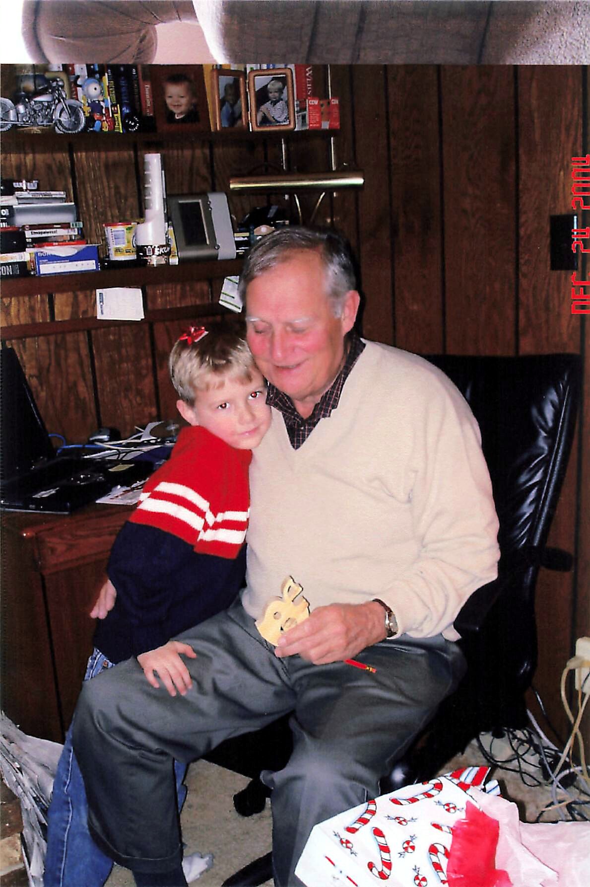 Grandfather and young boy share a warm hug in a cluttered home office, with presents and photos on wood-paneled walls nearby.