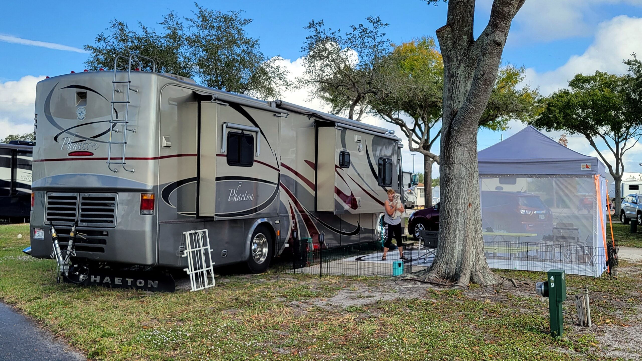 Large gray motorhome parked at a campground with a person holding a small dog near the slide-out and a blue tent canopy nearby.