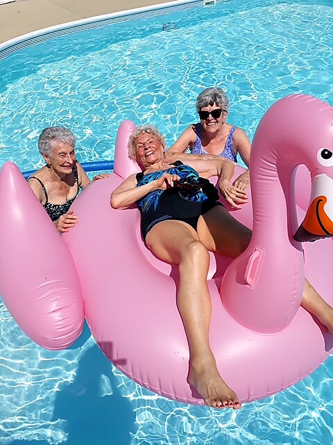Three smiling elderly women lounging on a pink inflatable flamingo in a sunny pool.