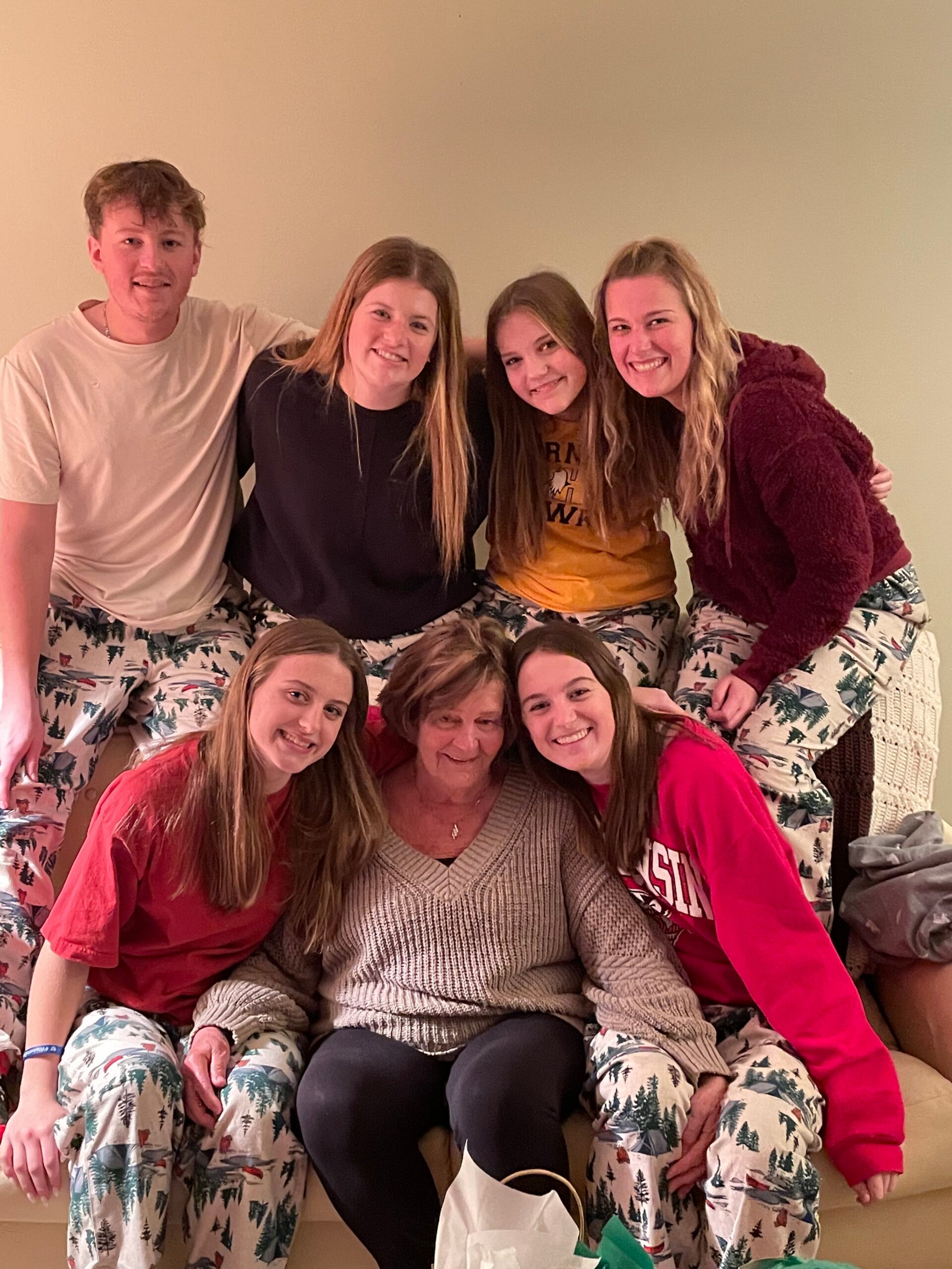 Group of eight people posing on a couch in matching pine-tree pajama pants, with a grandmother in a beige sweater at center, smiling.