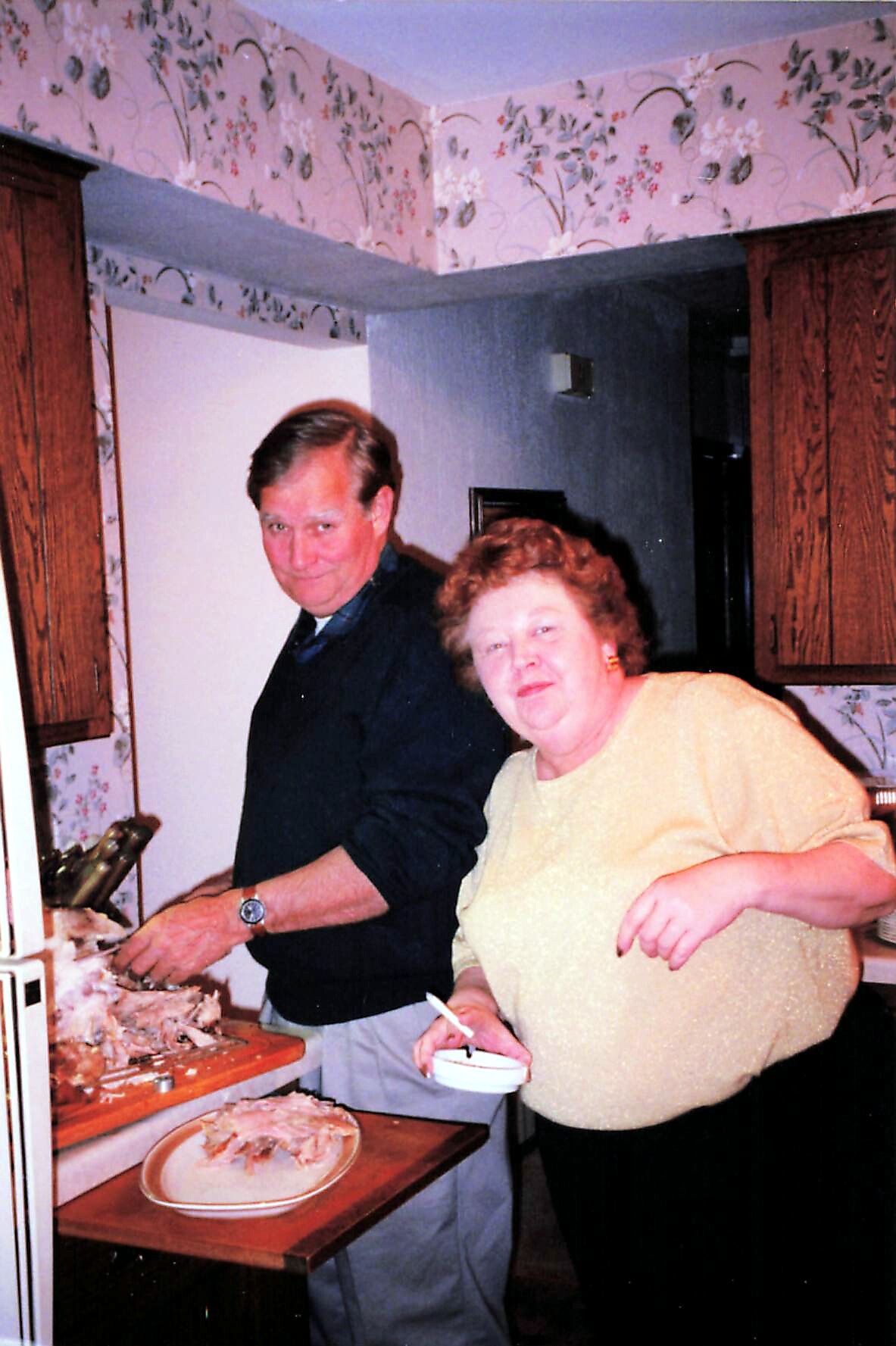 Man and woman carve turkey at a kitchen counter; plates of sliced meat are on the table in front.