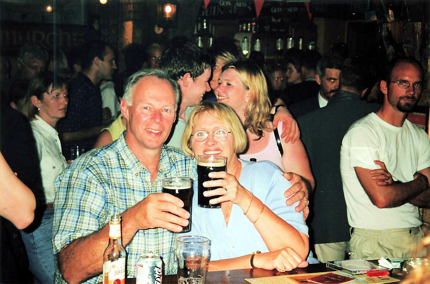 Smiling couple in the foreground raise beer glasses at a crowded bar, surrounded by friends partying in the background.