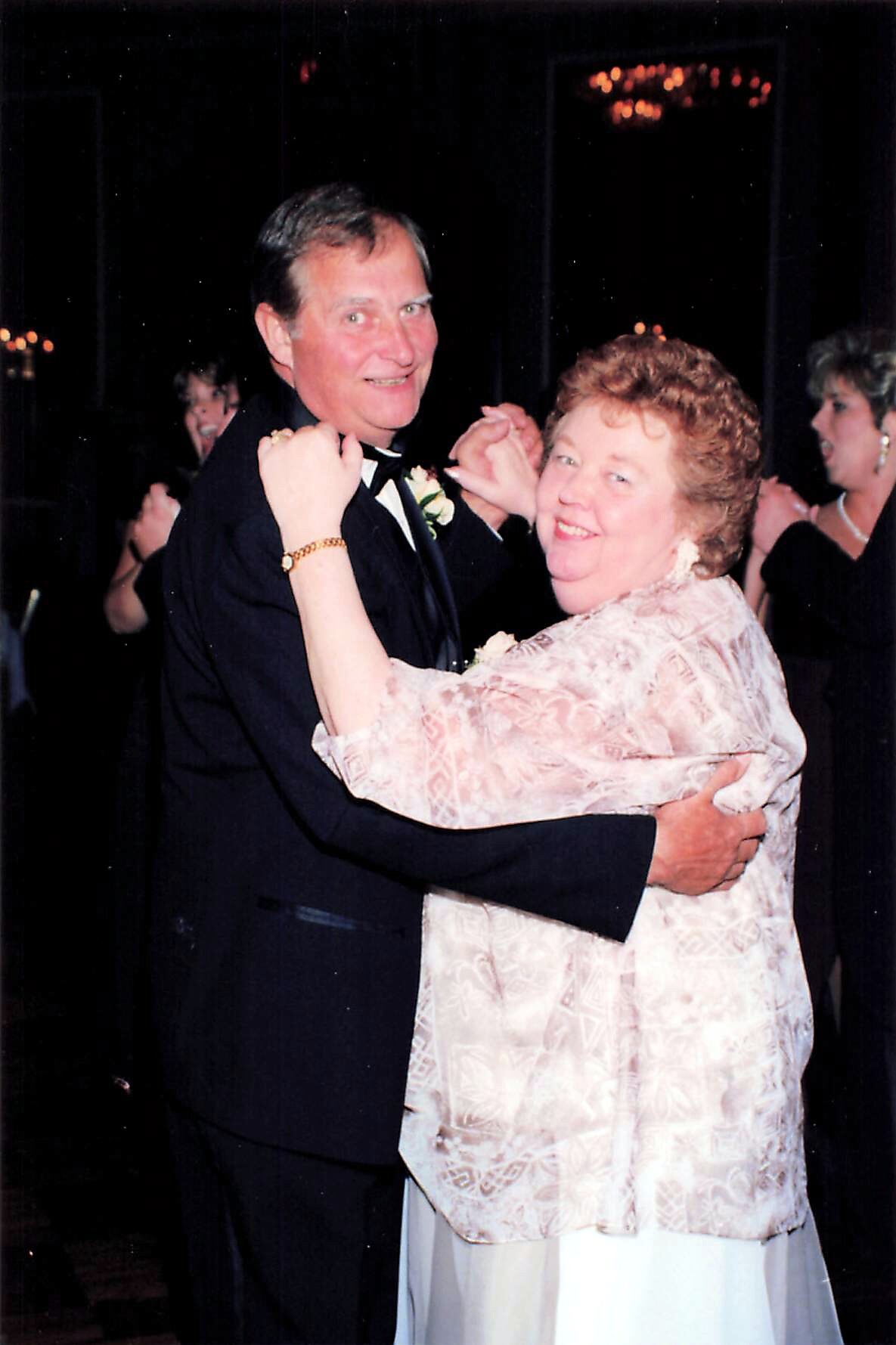 Older couple shares a dance at a formal event; man in a black tuxedo with boutonniere and a woman in a pale pink patterned jacket smiling at the camera.