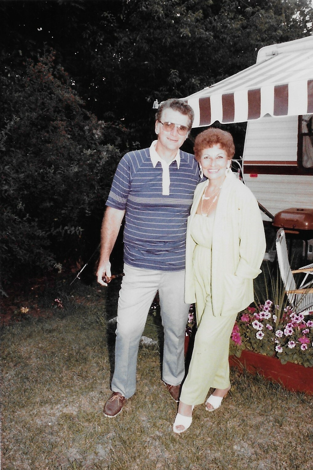 A smiling couple posing outdoors beside a striped camper while standing on the grass; man in a blue striped polo and jeans, woman in a lime-green outfit and white sandals.
