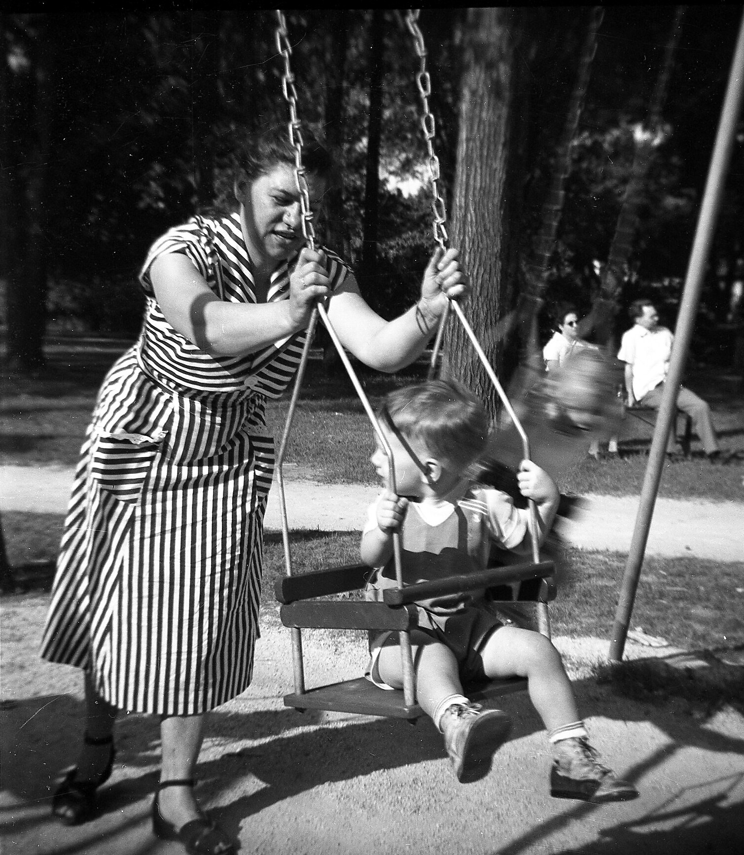 Woman in a striped dress helping a child on a swing in a park, chains and swing set visible; other people in background.