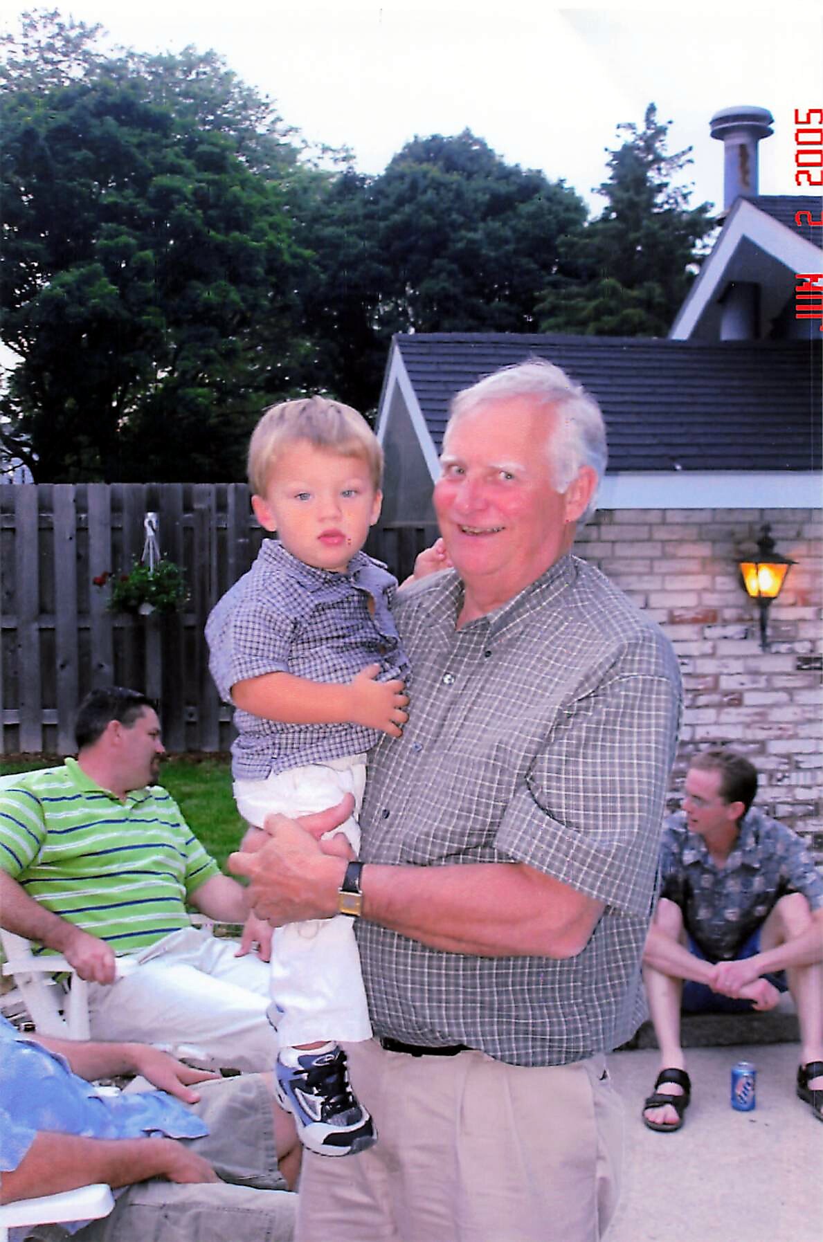 Older man in a checked shirt holding a young child at a backyard gathering, with other guests seated in the background and a brick wall behind them.