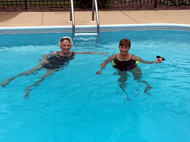 Two older women in a bright blue pool near the ladder, smiling; one wears sunglasses on her head, the other holds a small device.