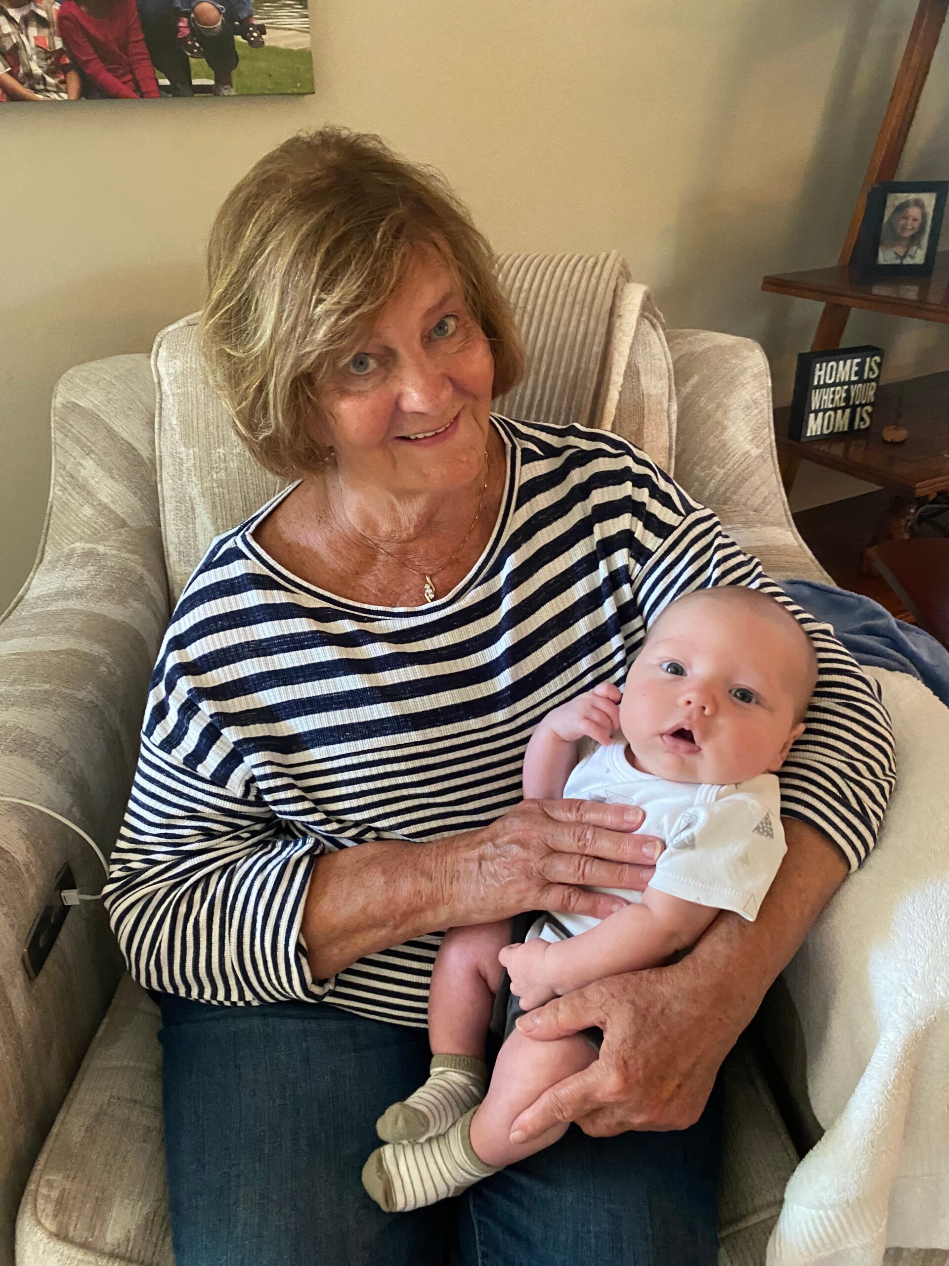 Older woman with short hair in a striped shirt cradles a newborn baby on her lap while sitting in a beige armchair at home to create a family moment.