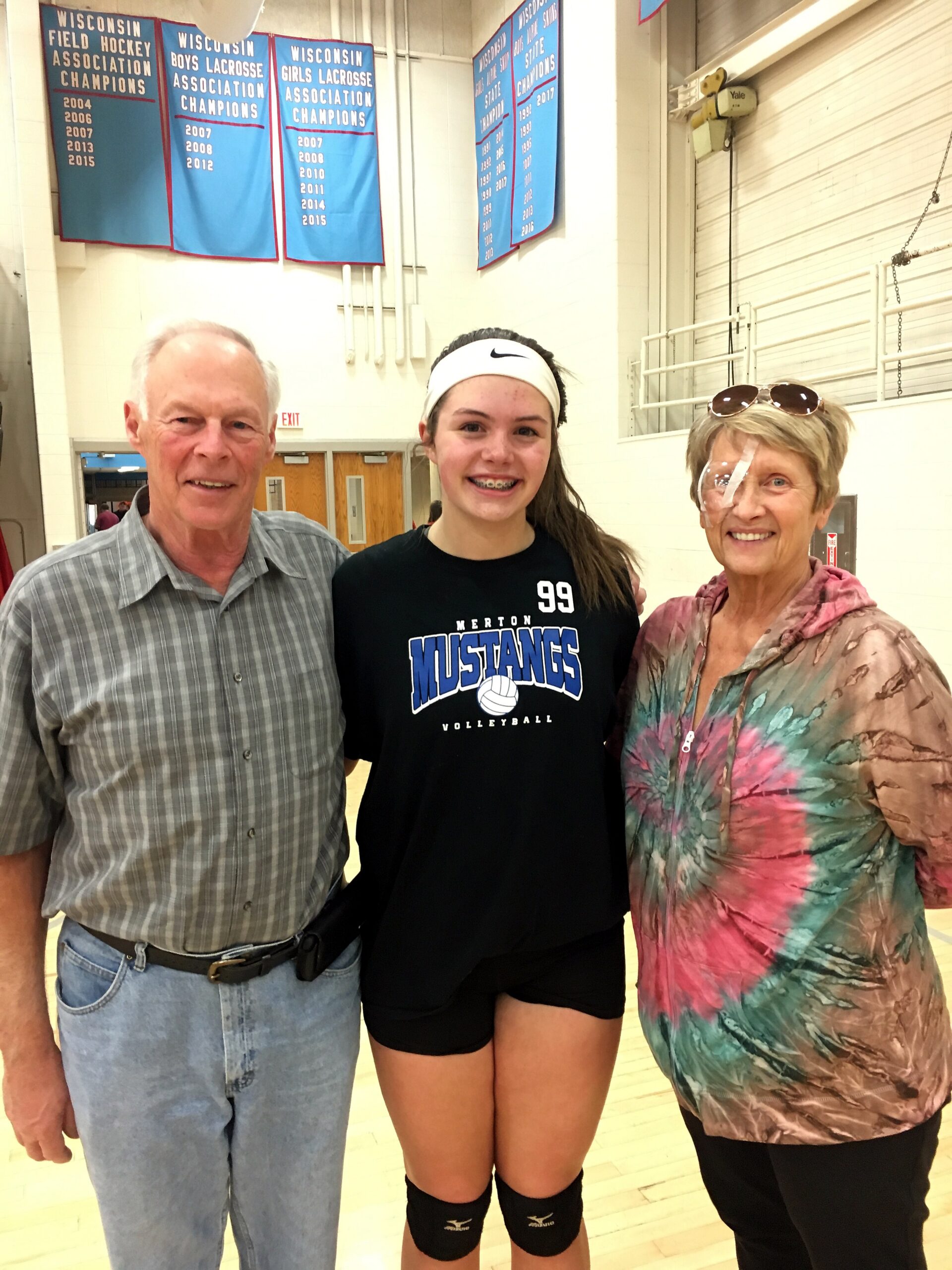 Teenage girl in a volleyball uniform and knee pads stands beside her smiling parents in a school gym with blue banners on the wall behind them.