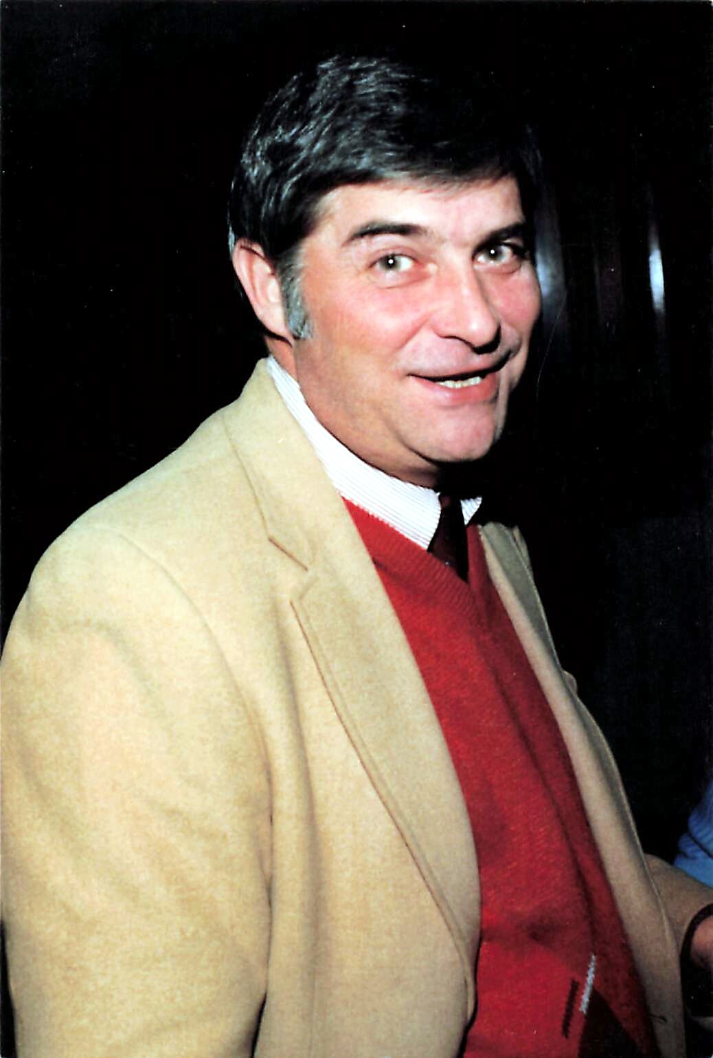 Smiling man with dark hair wearing a beige blazer over a red sweater and white striped shirt, posing for a photo.