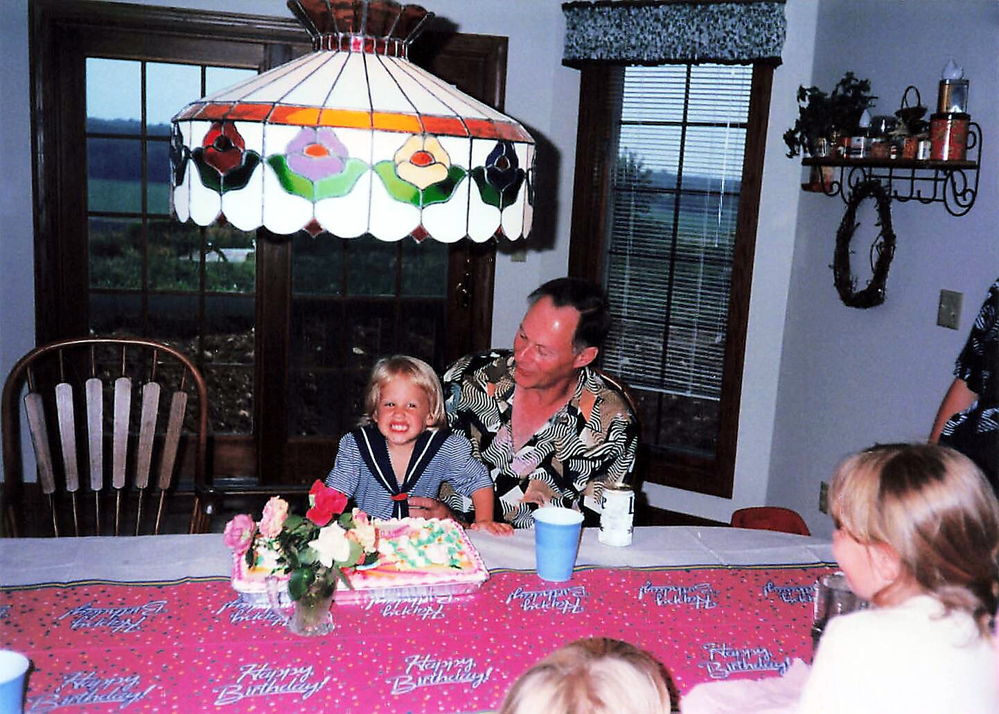 Young girl in a sailor dress smiles at a birthday cake on a pink tablecloth with 'Happy Birthday' text, seated beside a man in a floral shirt.