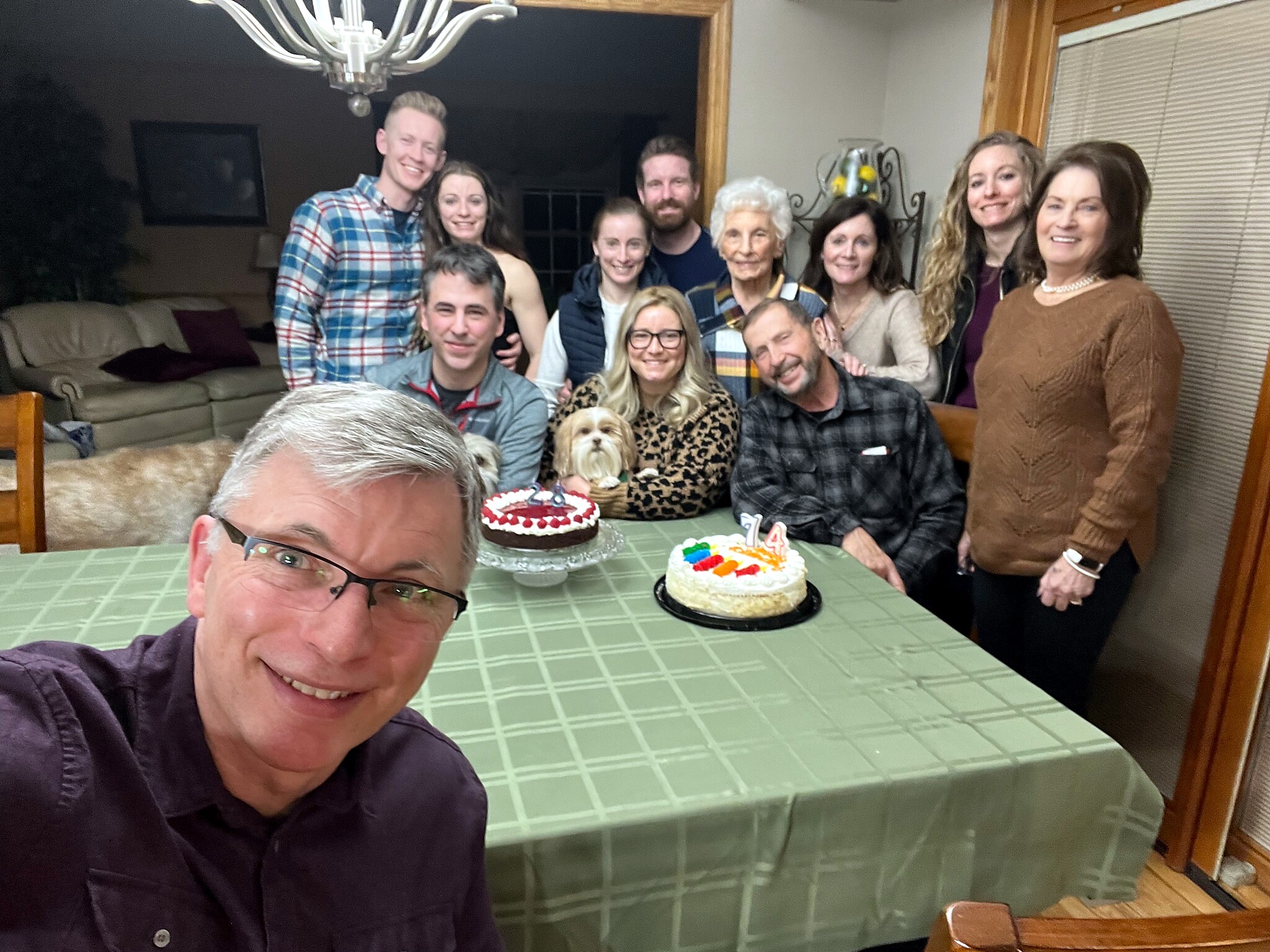 A group of friends and family gather around a kitchen table for a birthday, with the man in the foreground taking a selfie as two decorated cakes sit on the table and a small dog visible among the people.
