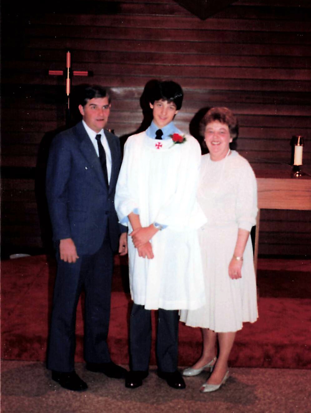 Young person in a white ceremonial gown with a red boutonniere stands between a man in a suit and a smiling woman in a light dress inside a church.