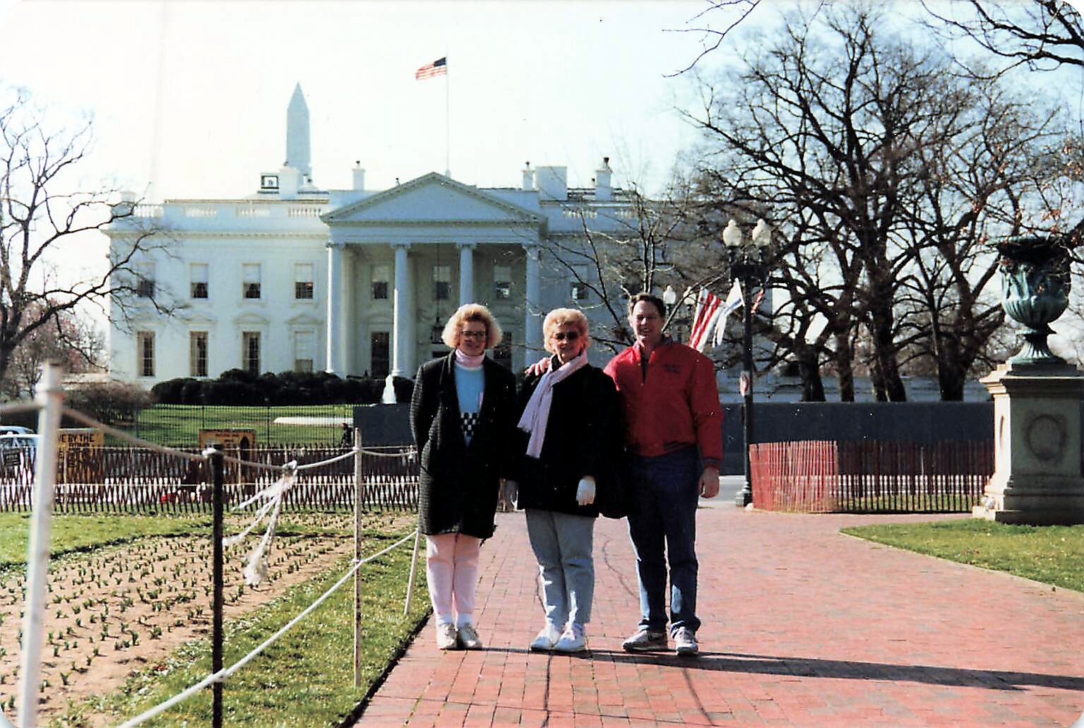 Three people pose for a photo on a brick walkway in front of the White House, with an American flag overhead and leafless trees nearby.