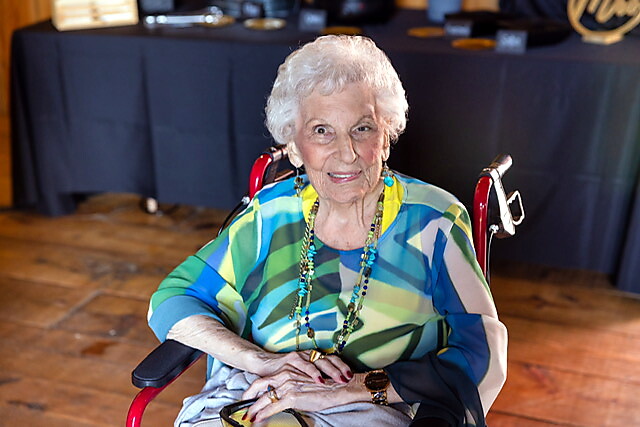 Elderly woman in a colorful blouse seated in a wheelchair at a social event, smiling at the camera.
