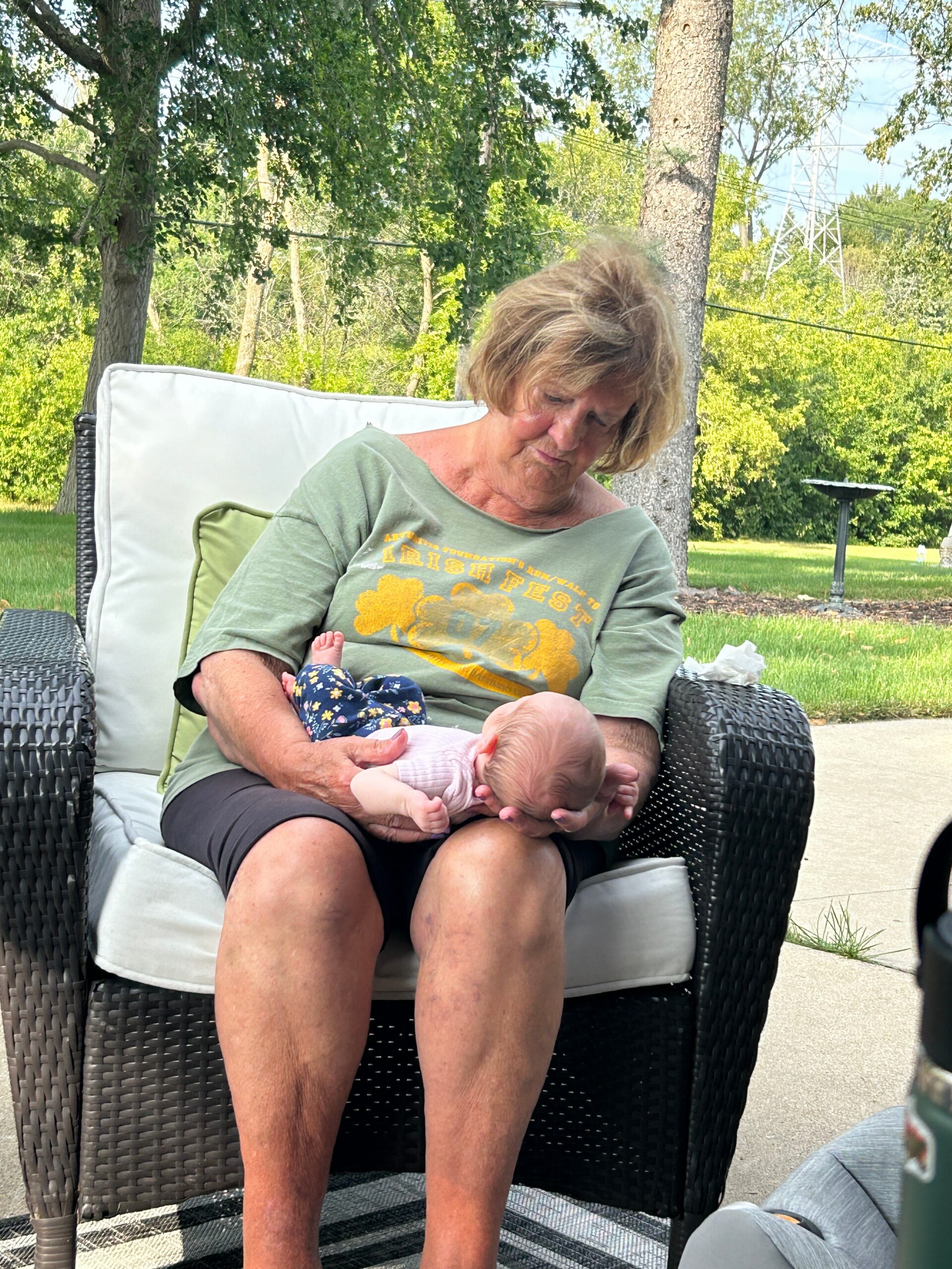Older woman sitting in a wicker chair outdoors, cradling a newborn baby in her arms.