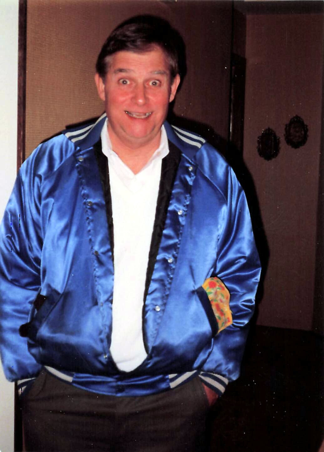 Man in a bright blue satin jacket and white shirt posing indoors, smiling at the camera.