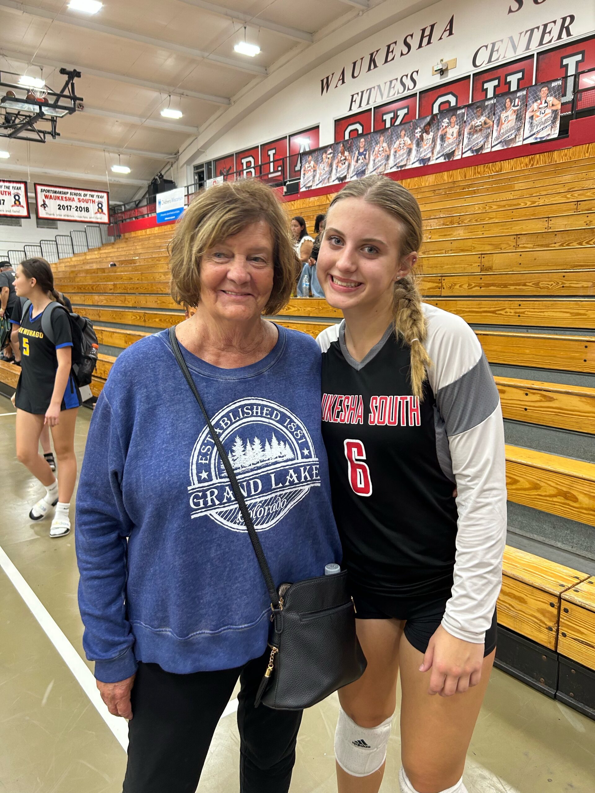 Smiling older woman in a blue sweatshirt posing with a Waukesha South volleyball player in a gym with wooden bleachers.