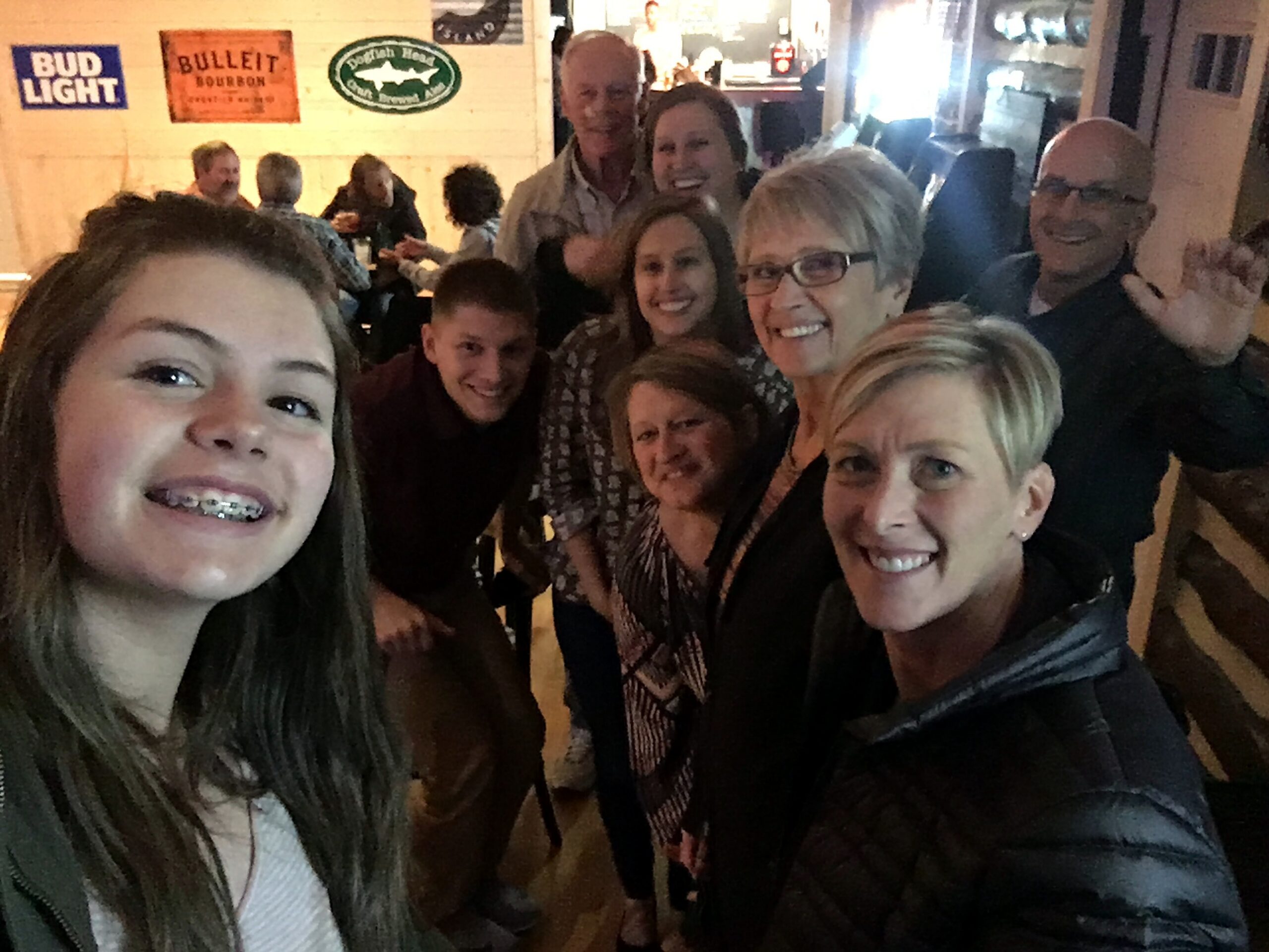 Smiling group selfie in a busy restaurant/bar, with friends gathered closely for the photo and wall signs in the background.