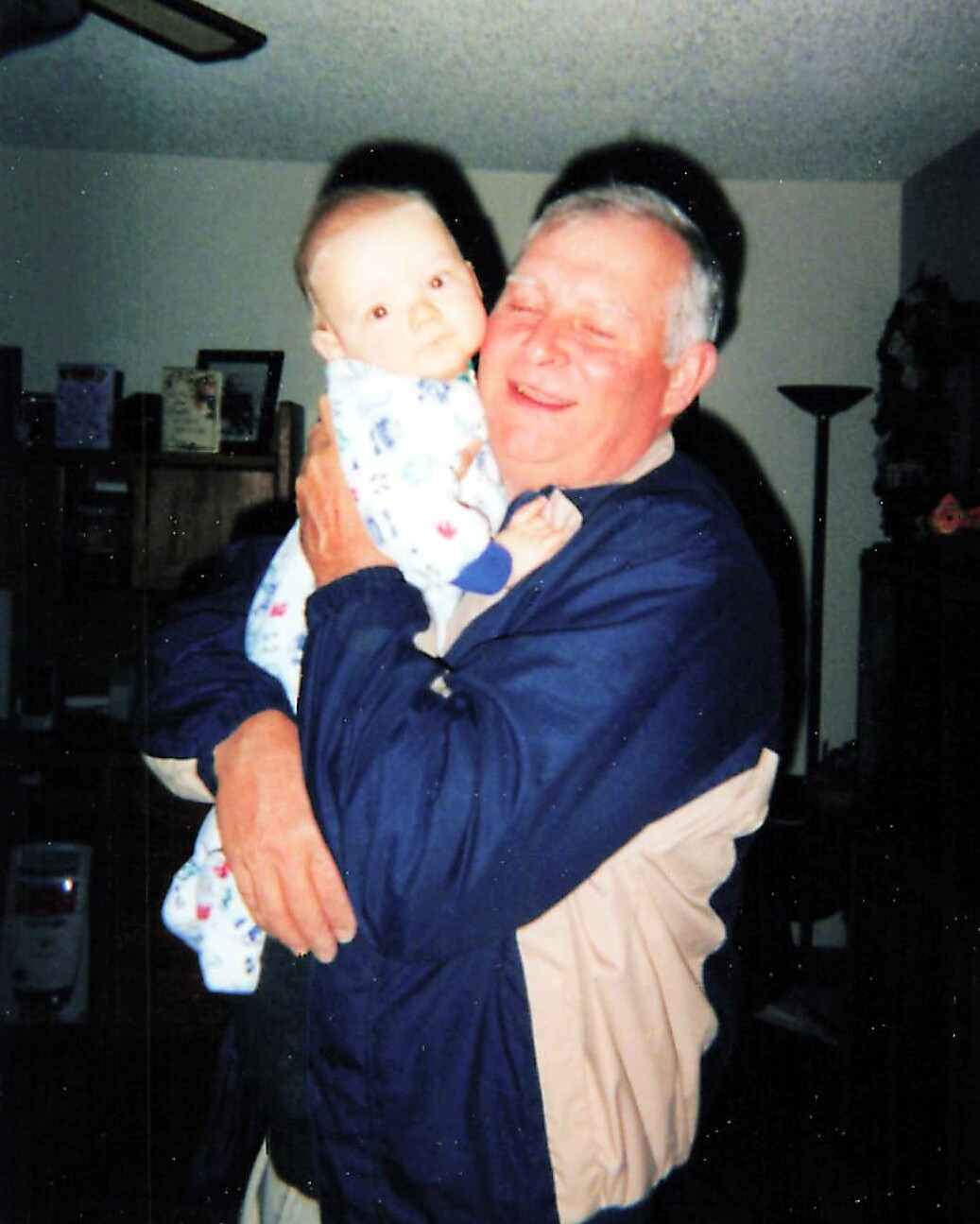 Older man in a blue jacket tenderly holds a baby wrapped in a pastel blanket indoors, both smiling softly at the camera