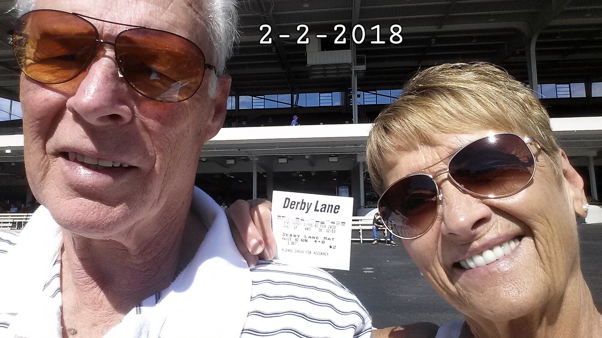 Close-up selfie of an older man and woman wearing sunglasses, holding a Derby Lane race ticket in front of a racetrack.