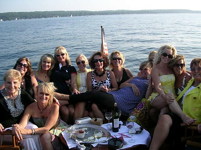 Group of women on a boat cruise by a lake, posing for a photo with drinks on a table in front of them.