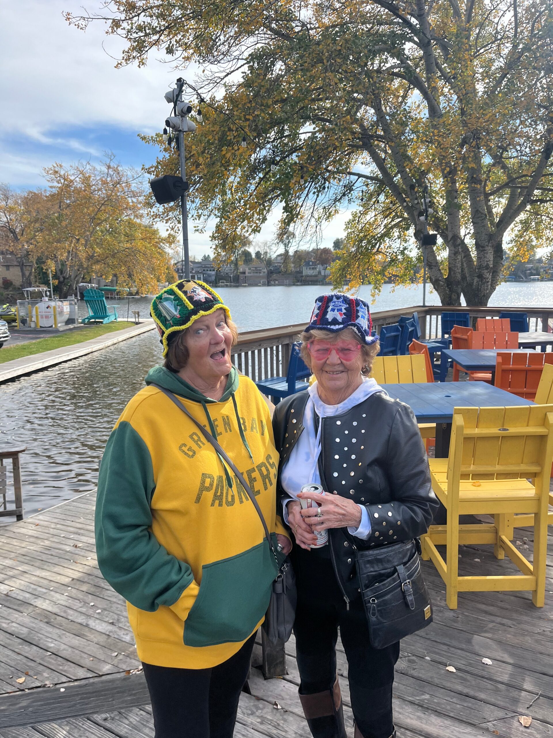 Two women pose on a wooden dock by the lake, wearing colorful hats and casual jackets, smiling at the camera with fall trees in the background.