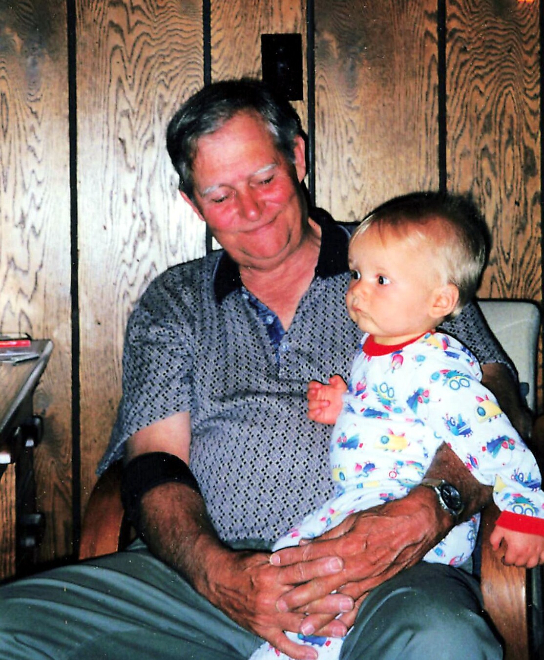 An older man smiles softly while holding a small child on his lap in a wood-paneled room.