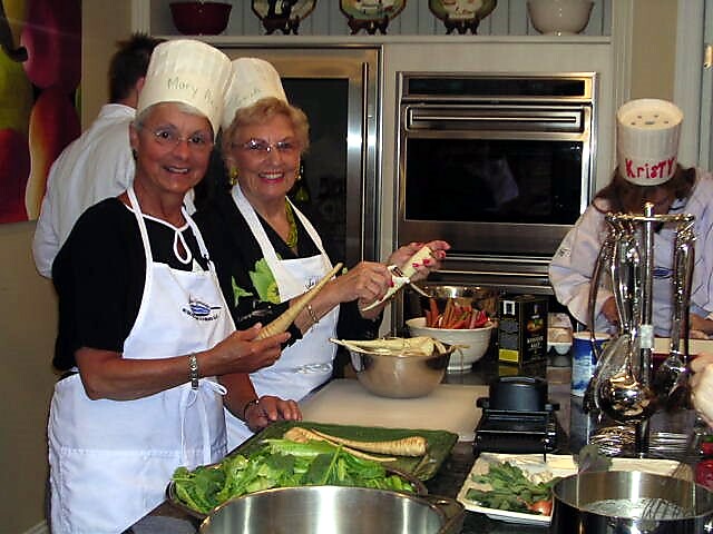 Two smiling women in chef hats and aprons demo cooking at a kitchen counter with greens and bowls in front of stainless steel appliances in the background.
