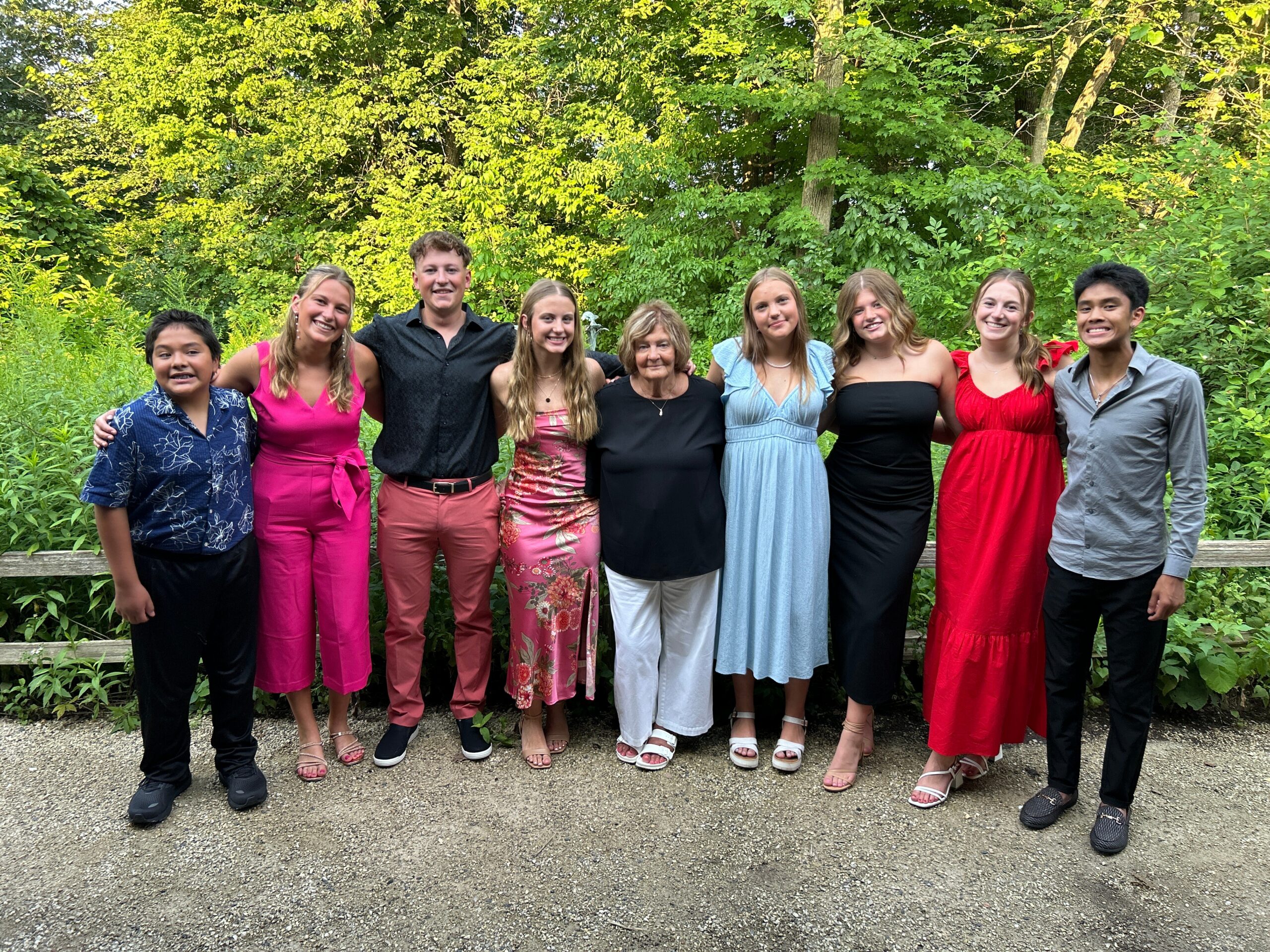 Nine people stand together with arms around each other in a sunny, green garden setting, smiling at the camera.