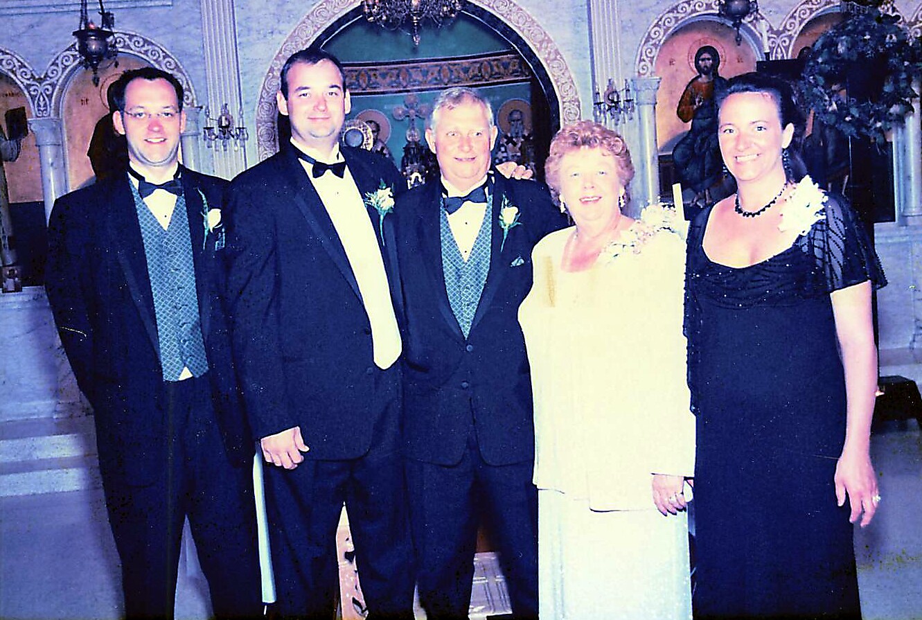 Group of five adults at a formal event in a church, with three men in tuxedos and two women in evening gowns posing for a photo.