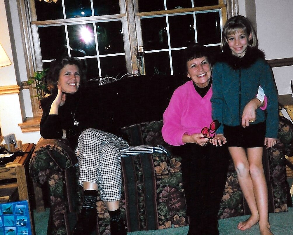 Three people posing on a floral sofa in a living room; a woman in pink sits center with a girl in teal standing beside her.