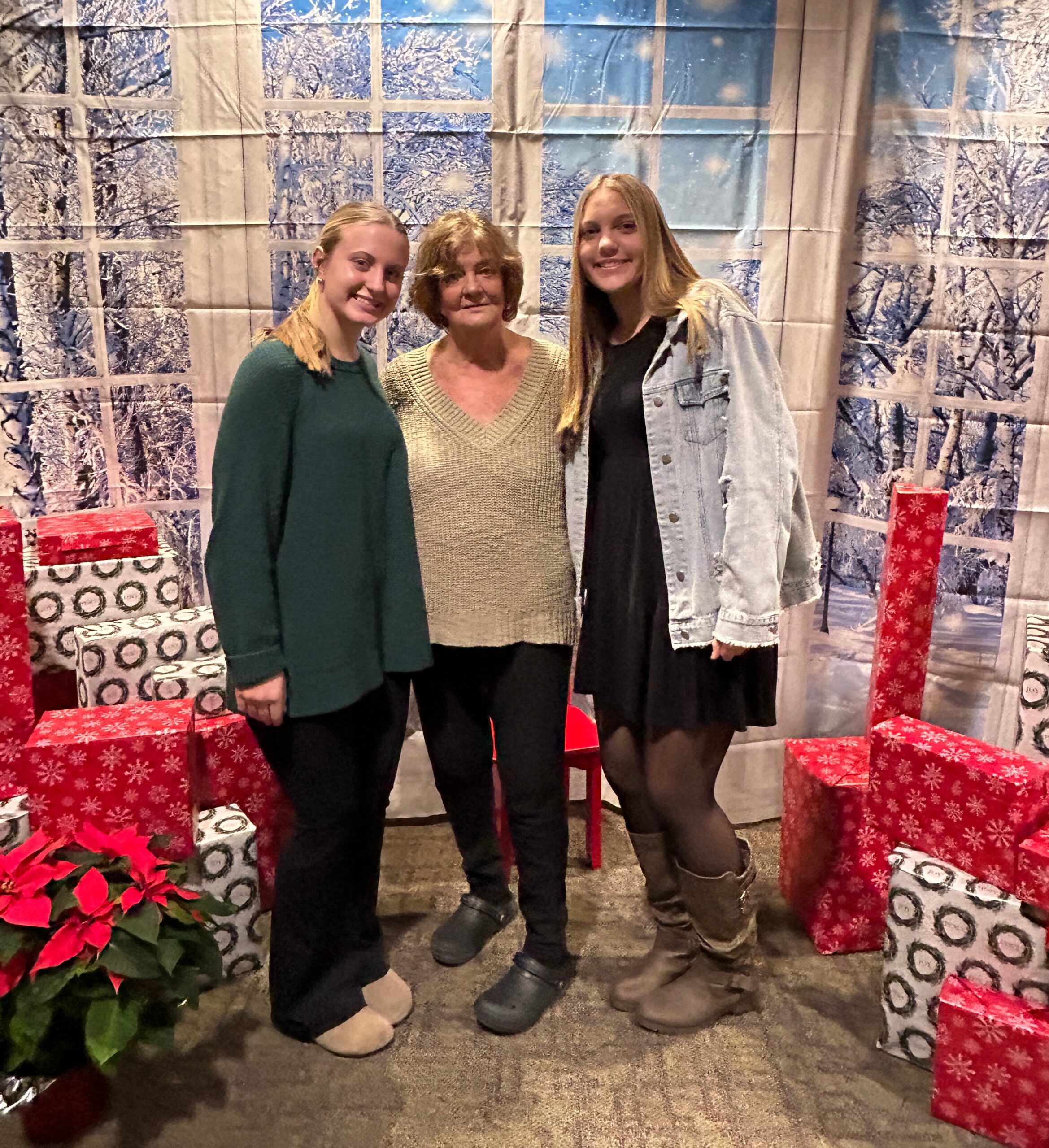 Three women posing together in front of a snowy, windowed backdrop with wrapped Christmas gifts around them.