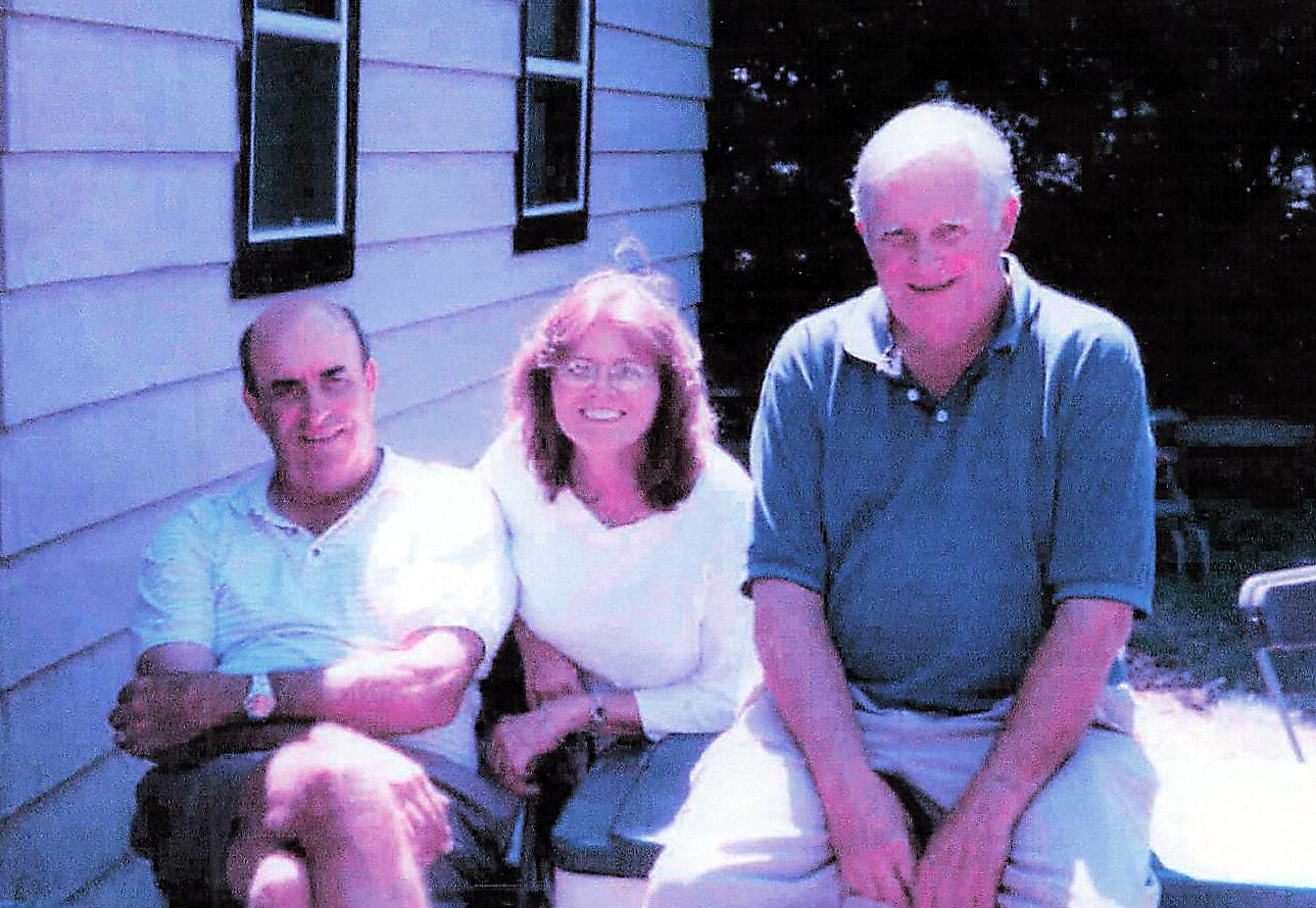 Three adults sitting together outside a light-colored house, smiling at the camera.