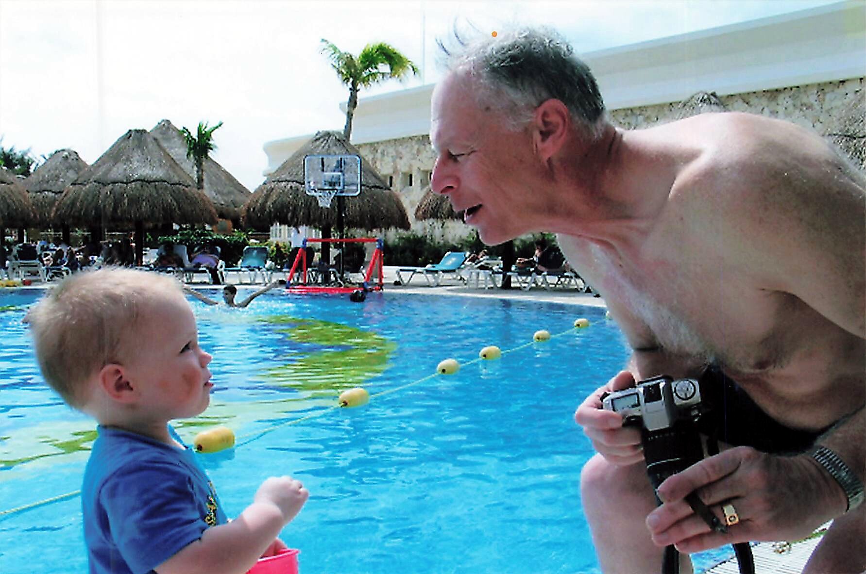 Man crouches by the pool edge, showing a camera to a young child while they face each other at a resort pool.