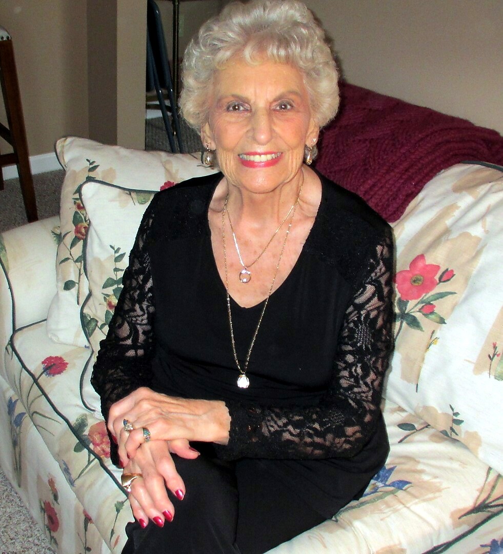 Smiling elderly woman with short white hair, wearing a black lace top and gold jewelry, seated on a floral-patterned sofa.