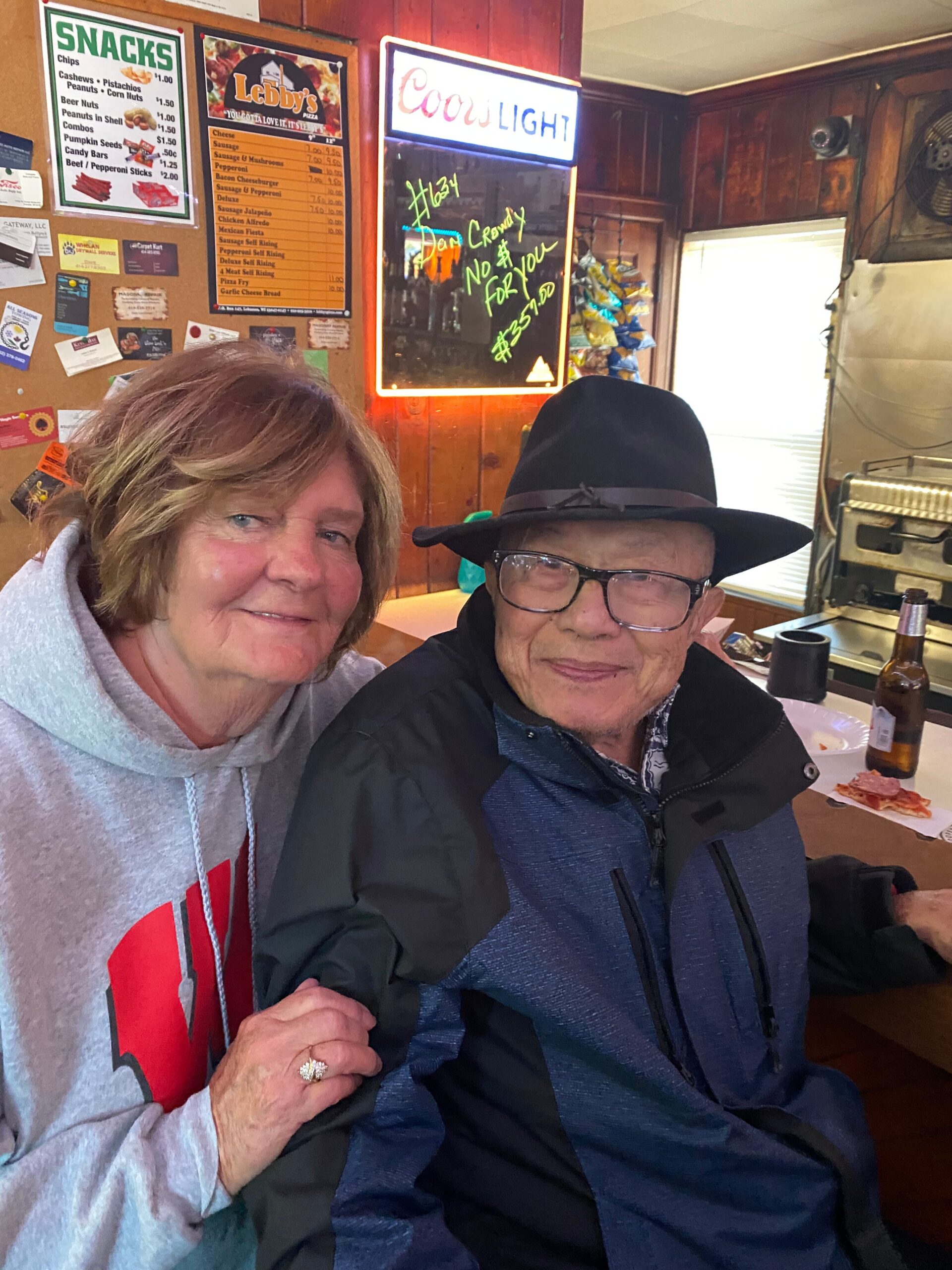 Two smiling seniors posing together at a wood-paneled diner counter, with snack menu and Coors Light neon sign in the background.