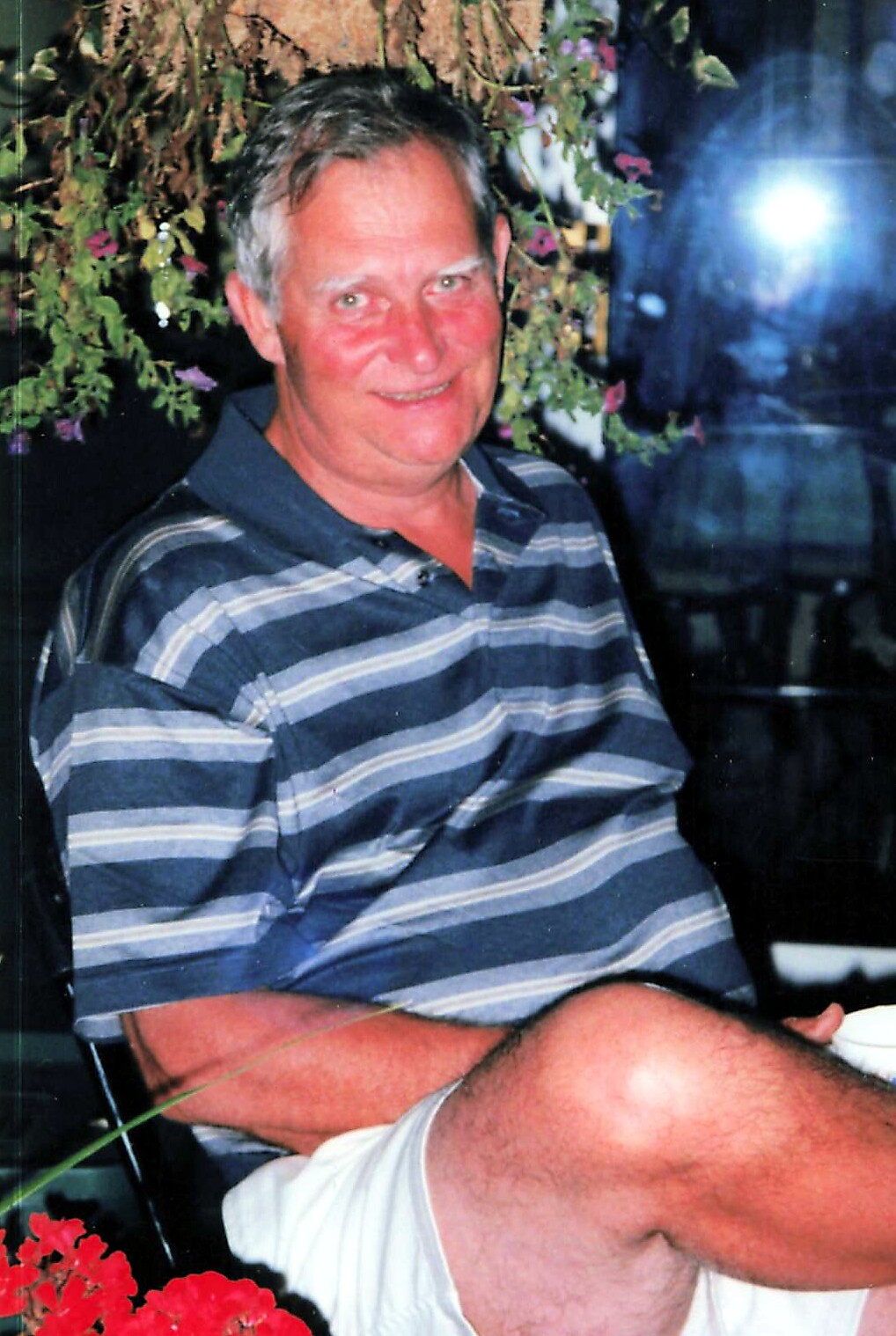 Older man with gray hair in a blue-gray striped polo shirt, smiling at the camera, seated outdoors among greenery and red flowers.