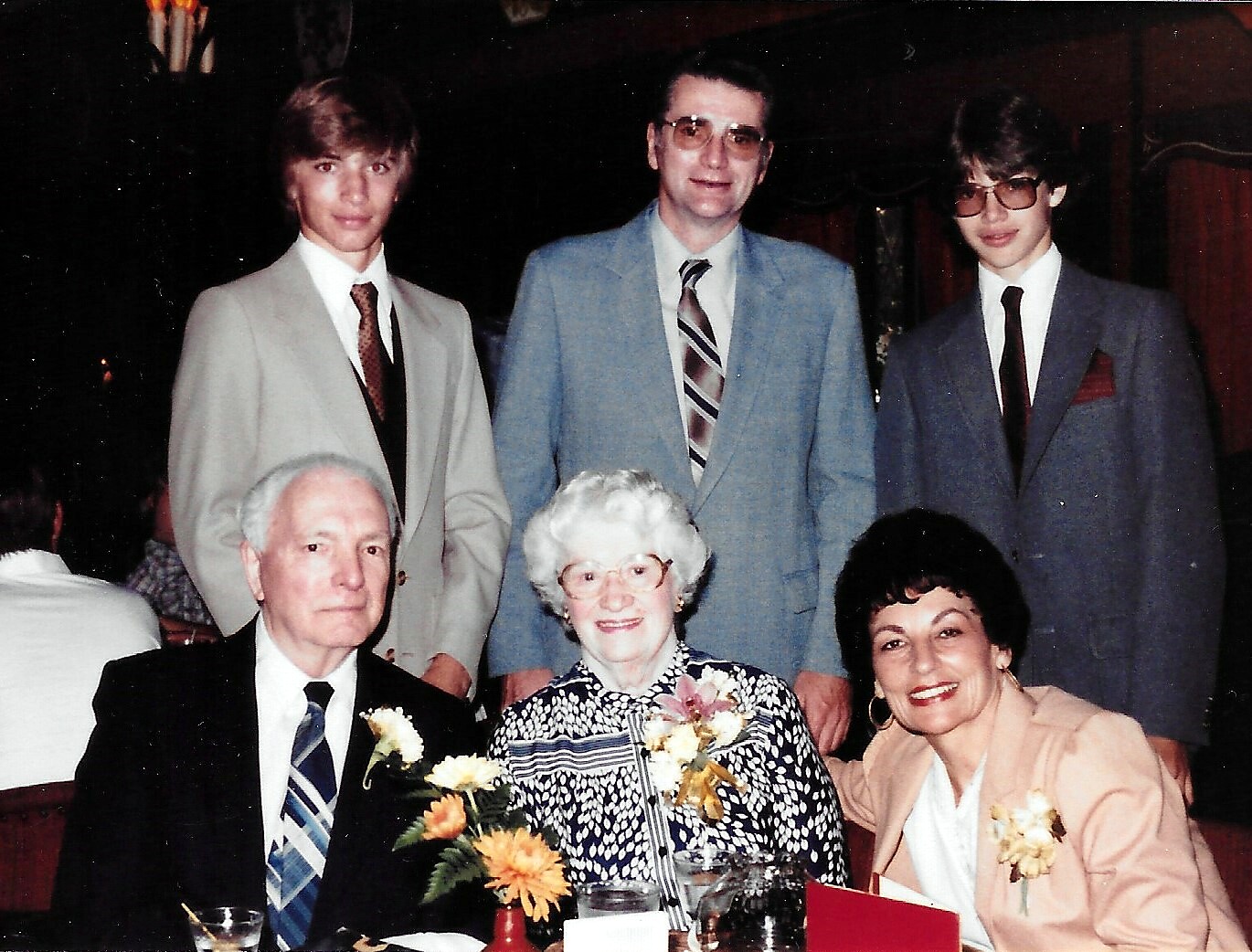 Six people in formal attire pose for a group photo at a banquet table, with floral arrangements in front and a dark background behind them.