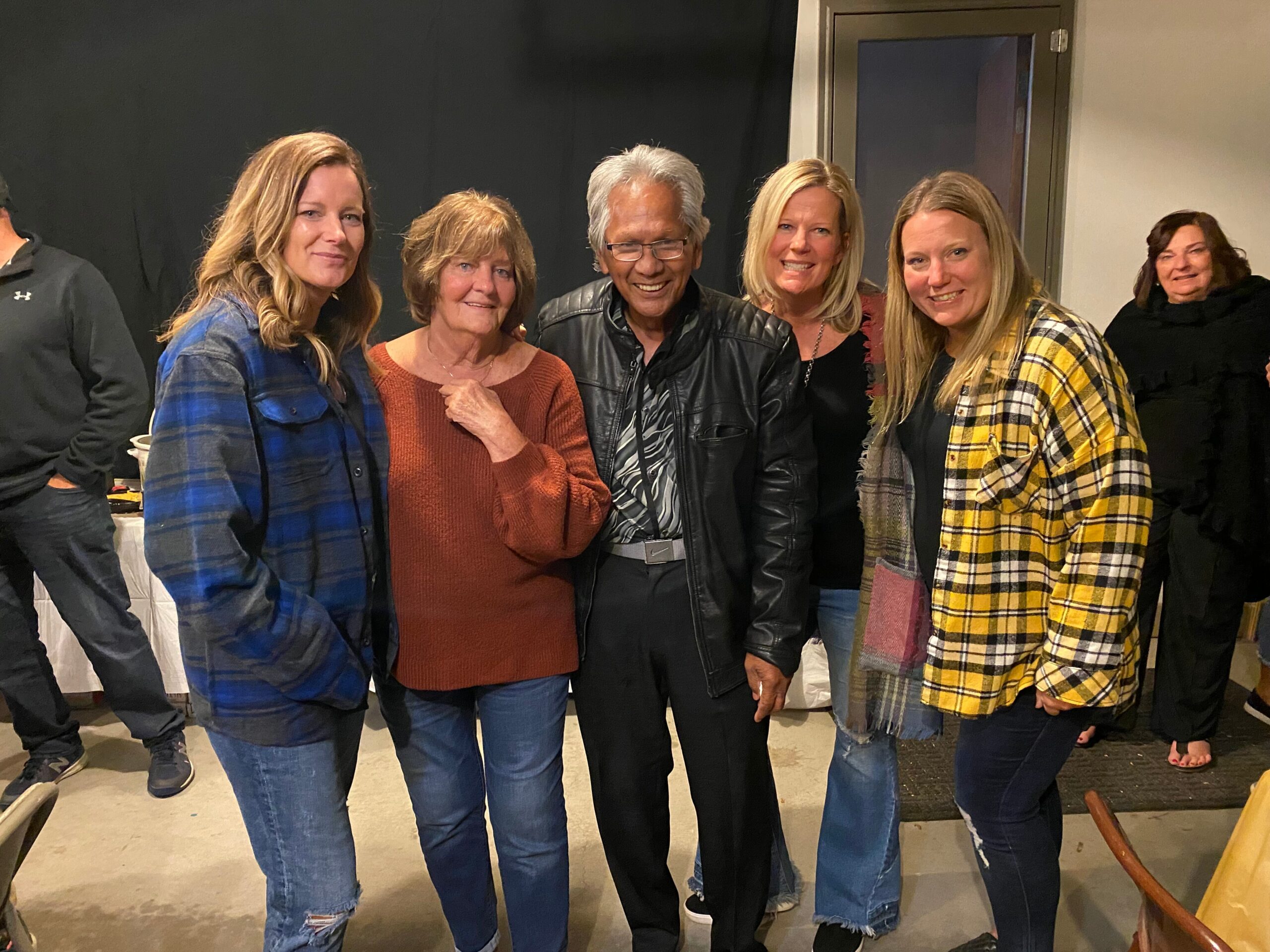 Group of seven adults posing indoors for a casual photo, smiling and wearing plaid shirts or jackets.
