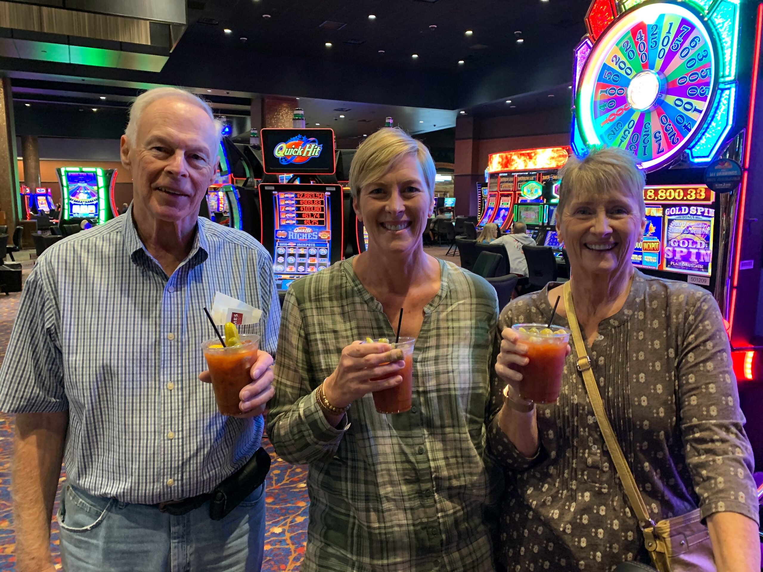 Three adults smiling, each holding a drink, in a casino with bright slot machines behind them.